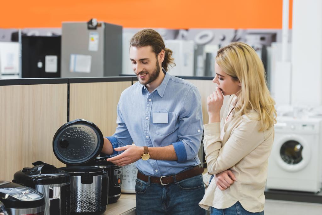 uśmiechnięty konsultant wskazujący ręką na powolną kuchenkę i zamyśloną kobietę patrzącą na nią w sklepie AGD  - Zdjęcie, obraz