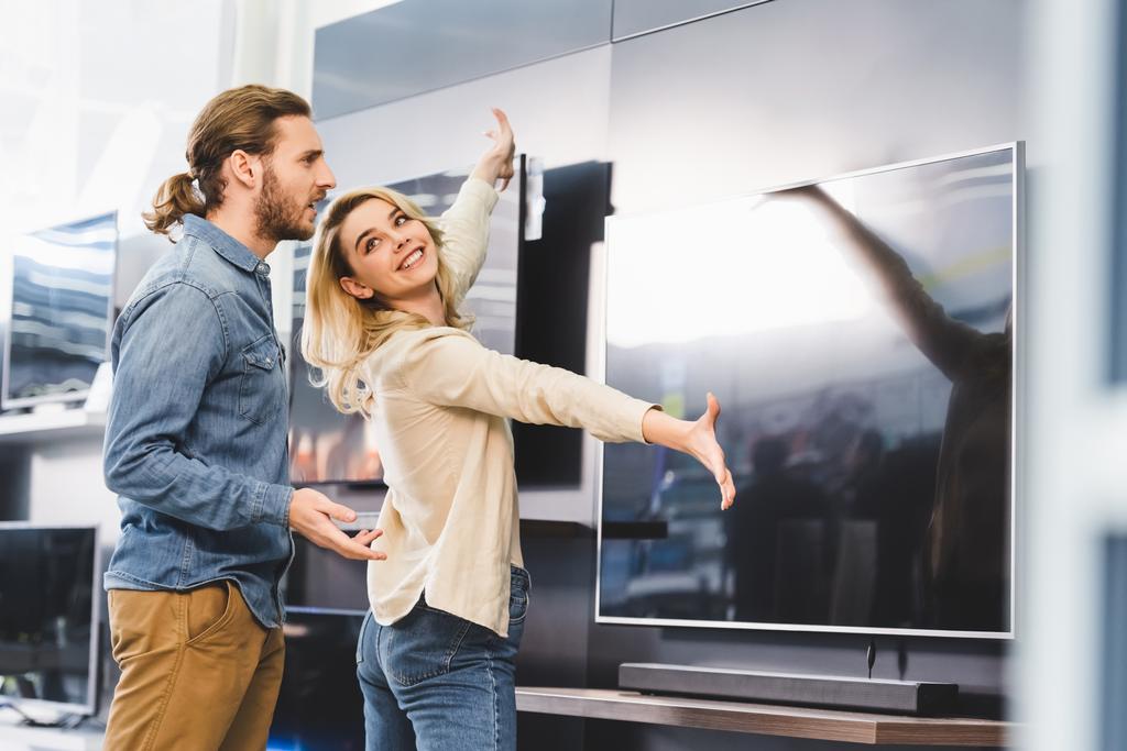 smiling girlfriend pointing with hands at tv and looking at shocked boyfriend in home appliance store  - Photo, Image