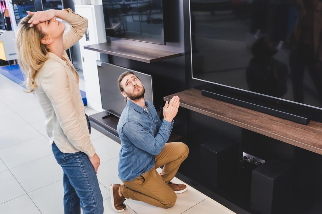přítel ukazuje prosím gesto v blízkosti televize a unavený přítelkyně dotýkání hlavy v obchodě domácí spotřebiče  - Fotografie, Obrázek