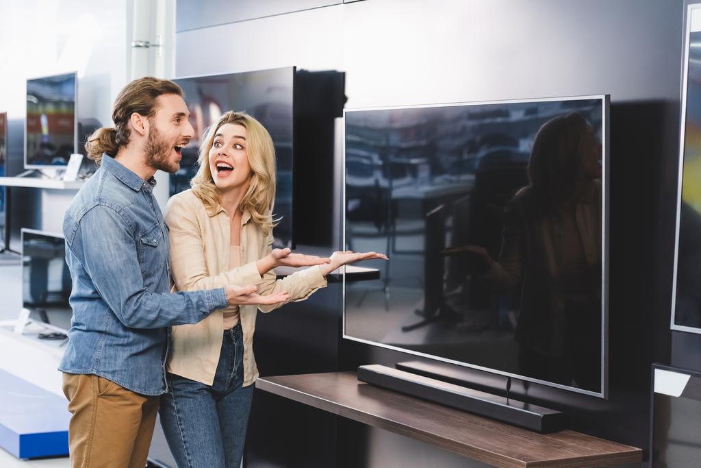 Schockierter Freund und Freundin zeigt mit Händen auf Fernseher in Haushaltsgerätegeschäft  - Foto, Bild