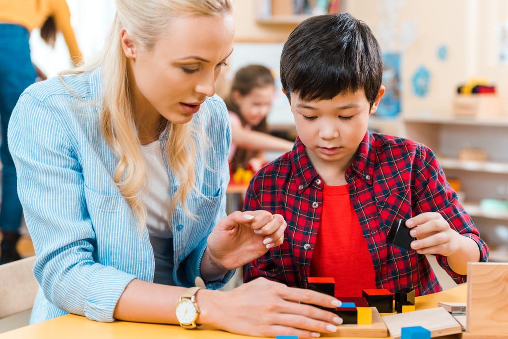 Foco seletivo de professor e criança jogando blocos de construção com crianças em segundo plano na escola montessori
 - Foto, Imagem