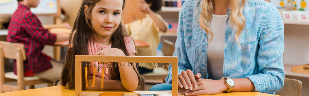 Селективный фокус ребенка, смотрящего на камеру во время игры учителя в школе Монтессори, панорамный снимок
 - Фото, изображение