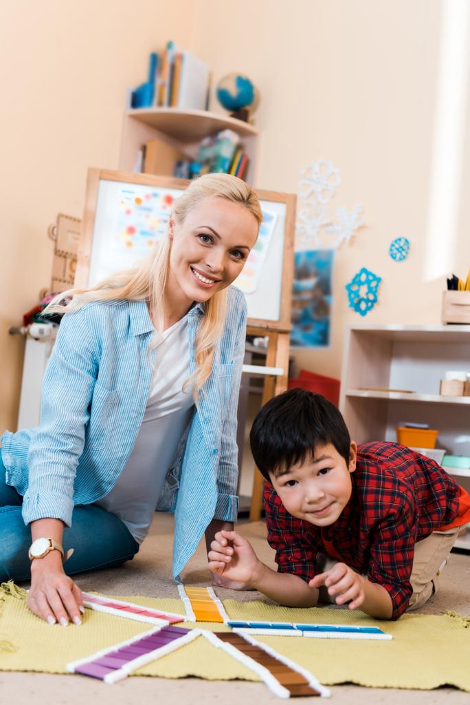 Επιλεκτική εστίαση του δασκάλου και του παιδιού χαμογελώντας στην κάμερα, ενώ παίζει το παιχνίδι στο πάτωμα στο σχολείο Montessori - Φωτογραφία, εικόνα