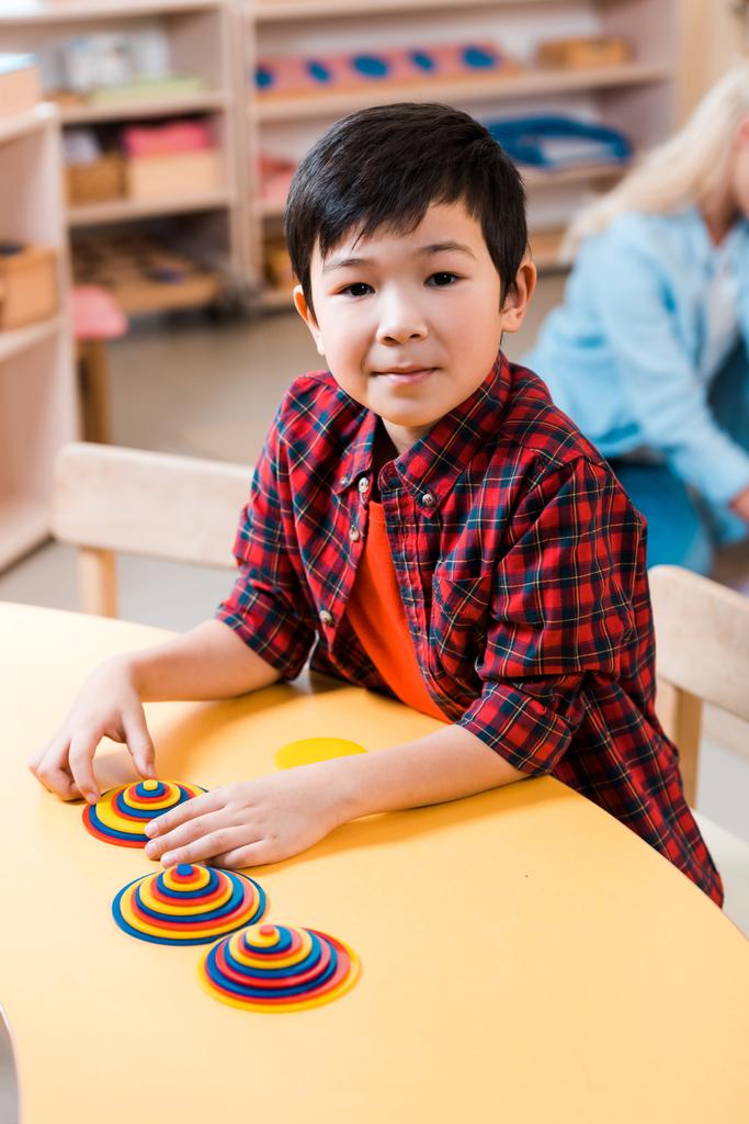 Selektywne skupienie azjatyckiego dziecka patrzącego w kamerę podczas zabawy za biurkiem w szkole montessori - Zdjęcie, obraz