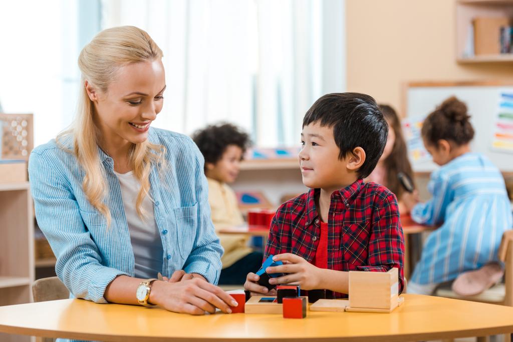Επιλεκτική εστίαση του χαμογελαστού δασκάλου και του παιδιού που παίζει δομικά στοιχεία με τα παιδιά στο παρασκήνιο στο σχολείο Montessori - Φωτογραφία, εικόνα