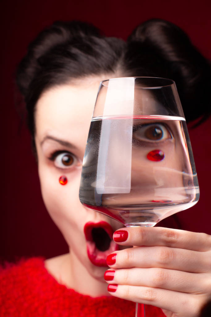 emotioneel meisje in rood met rode lippen en strass steentjes op haar gezicht houdt een glas in haar handen - Foto, afbeelding