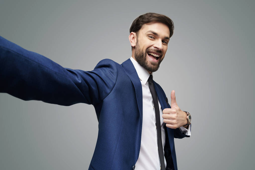 Weitwinkel-Selfie-Aufnahme eines jungen Geschäftsmannes, der auf grauem Hintergrund den Daumen nach oben zeigt - Foto, Bild