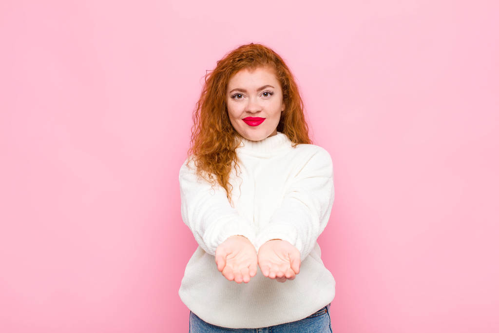 giovane donna testa rossa sorridente felicemente con amichevole, fiducioso, sguardo positivo, offrendo e mostrando un oggetto o un concetto contro muro rosa
 - Foto, immagini