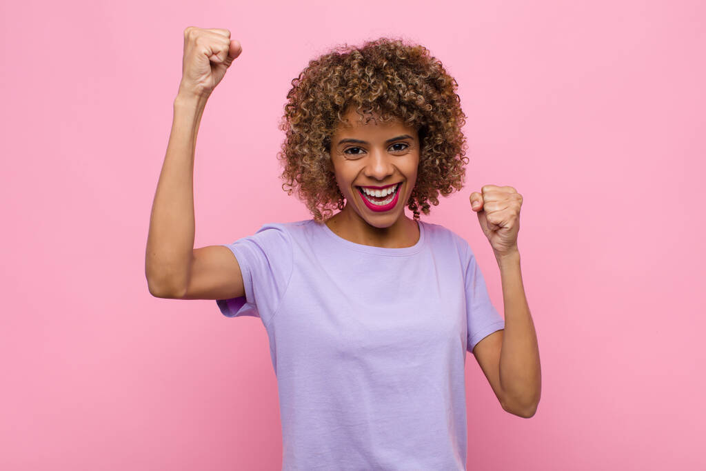 молодая африканская американка триумфально кричит, выглядит взволнованной, счастливой и удивленной победительницей, празднуя у розовой стены
 - Фото, изображение