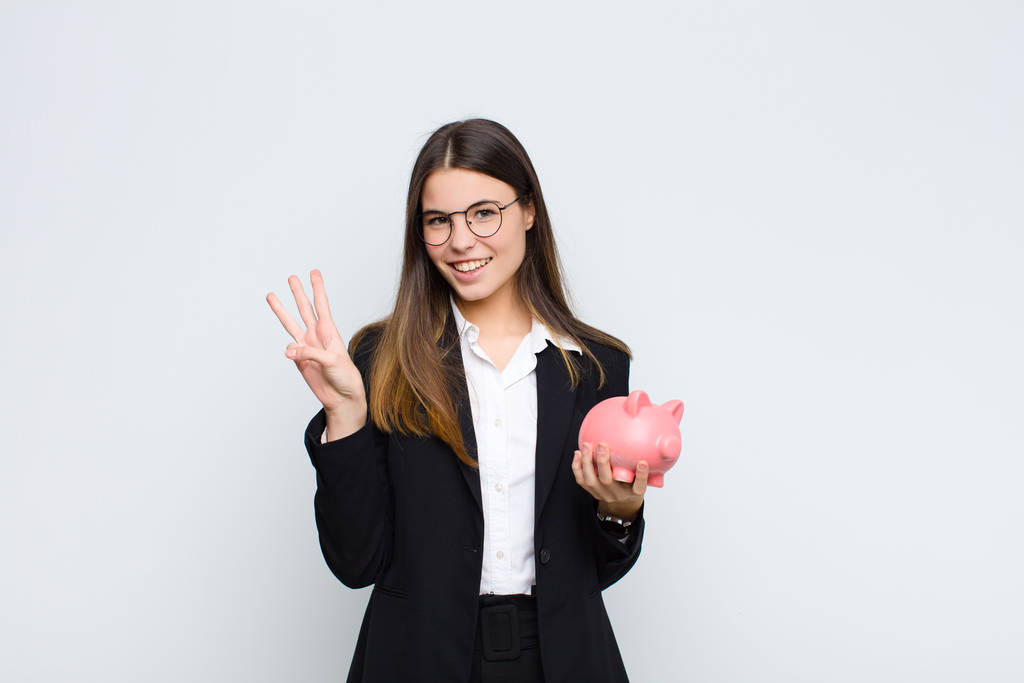young pretty woman smiling and looking friendly, showing number three or third with hand forward, counting down with a piggy bank - Photo, Image