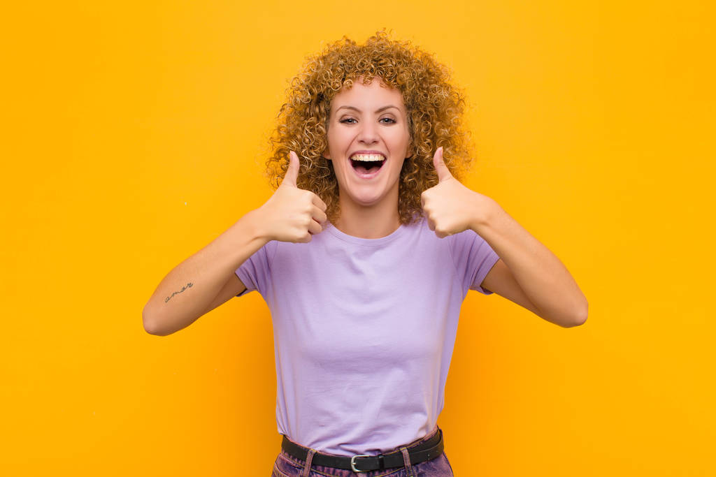 young afro woman smiling broadly looking happy, positive, confident and successful, with both thumbs up against orange wall - 写真・画像