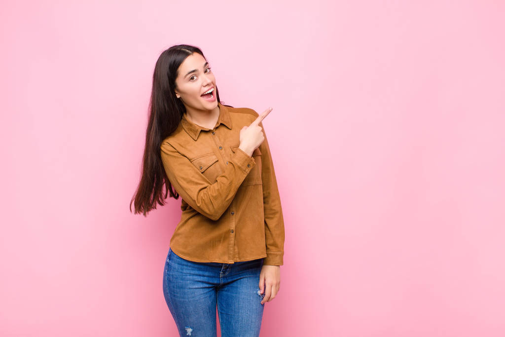 young pretty woman looking excited and surprised pointing to the side and upwards to copy space against pink wall - Photo, Image
