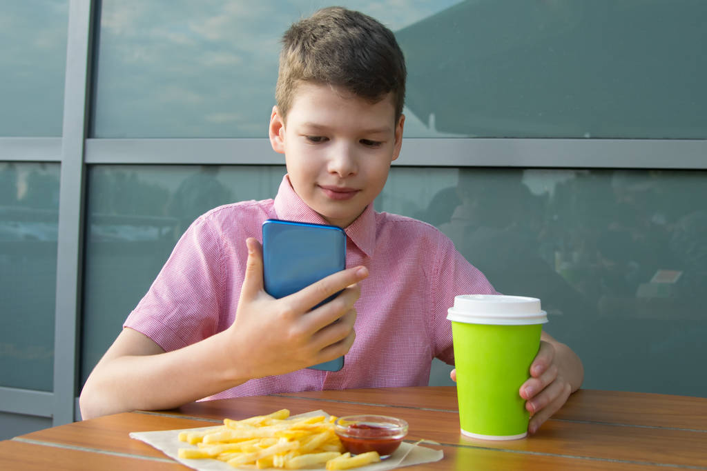 Der Junge sitzt am Tisch und isst Pommes mit Ketchup und einem Getränk, hält ein Telefon in der Hand - Foto, Bild