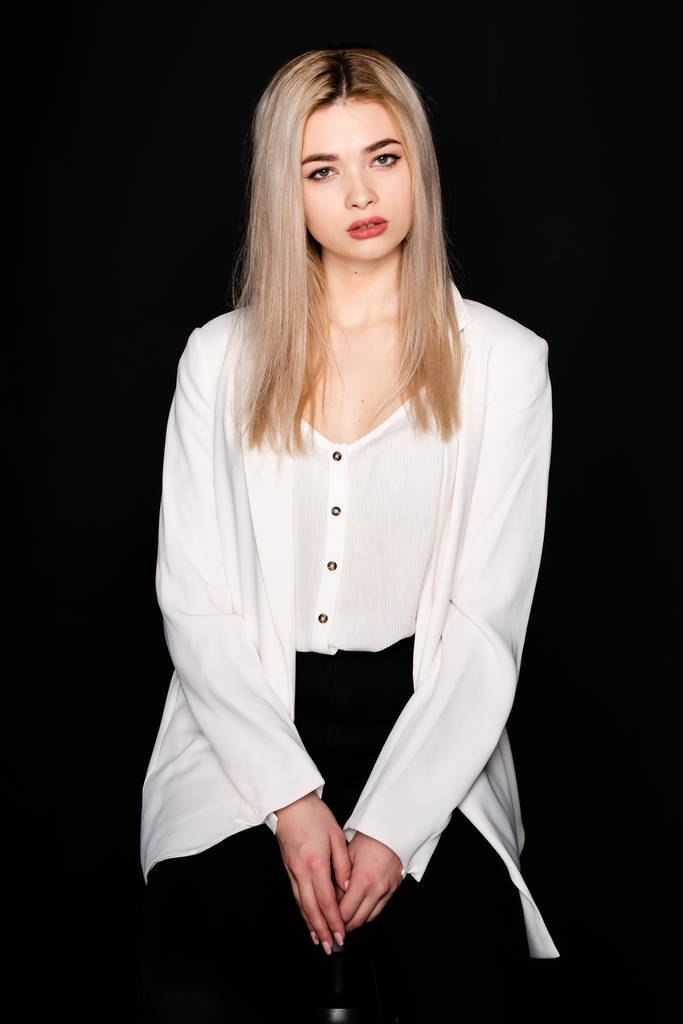 Young beautiful girl posing sitting on a chair in a white t-shirt and black trousers on a black background. Looks at the camera. - Photo, Image