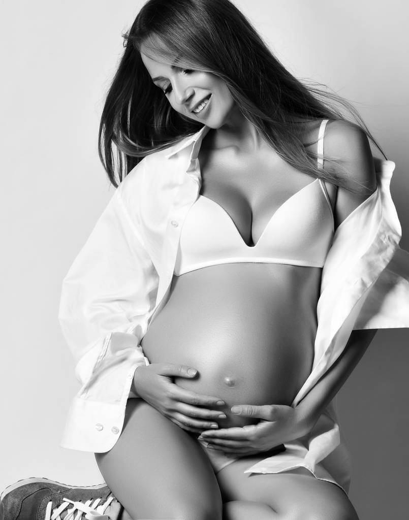 junge schöne schwangere Frau in weißem Hemd und Unterwäsche und Turnschuhen sitzend, den Bauch anschauend und lächelnd - Foto, Bild