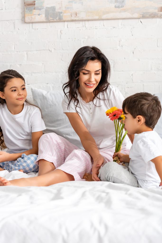 rozkošný chlapec drží květiny, zatímco sedí v posteli v blízkosti šťastný maminka a sestra na matky den - Fotografie, Obrázek