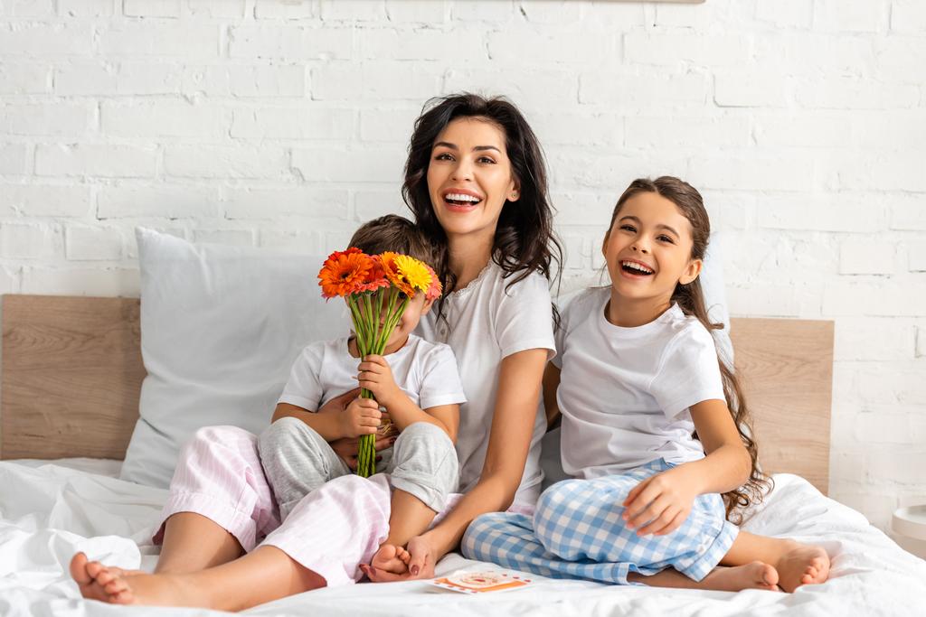 cute boy holding flowers while sitting in bed near happy mother and sister - Photo, Image