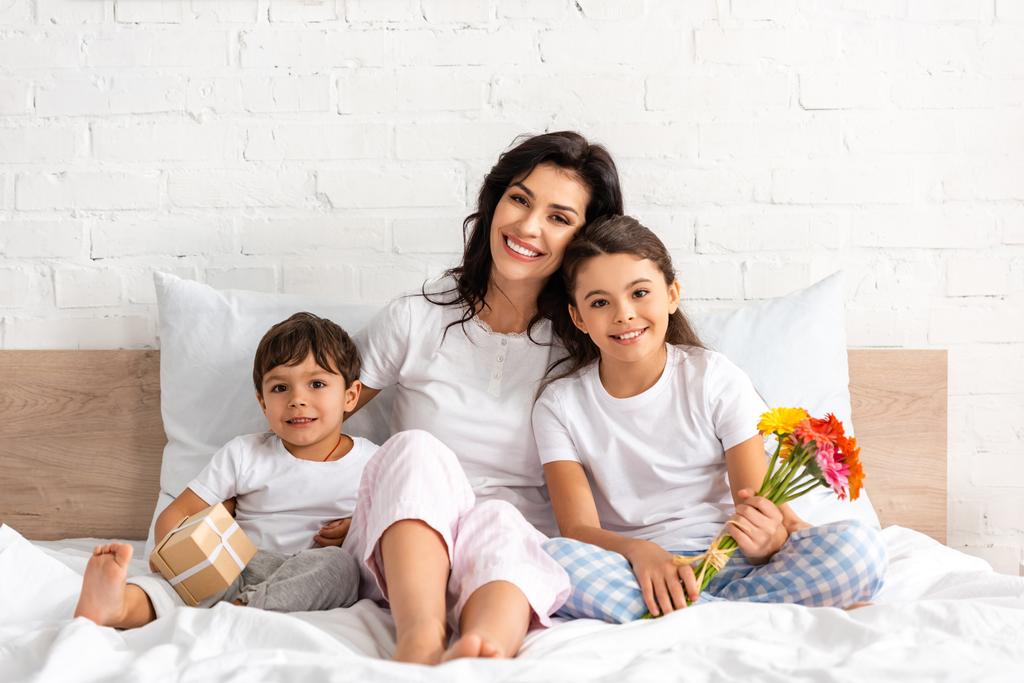 boldog nő gyerekekkel mosolyog a kamera előtt, miközben ül az ágyban virágokkal és ajándékdobozzal - Fotó, kép