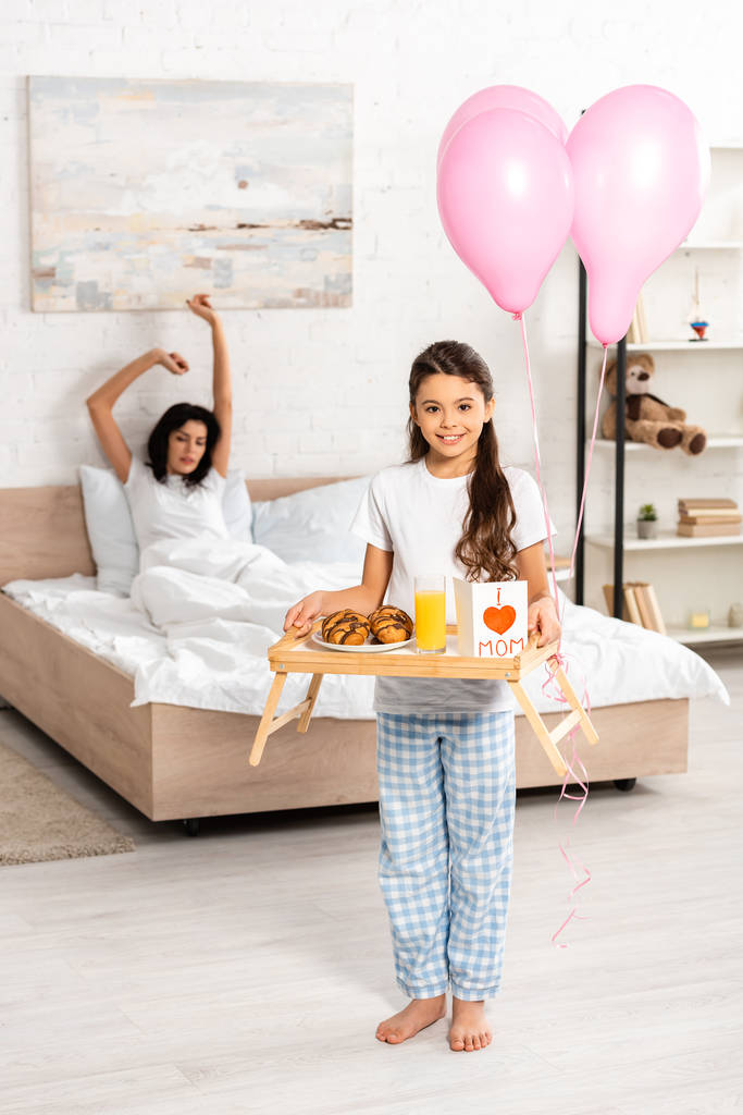 niedliche Tochter hält Tablett mit Frühstück, Muttertagskarte mit Herzzeichen und Mama Schriftzug, während Mutter sich im Bett dehnt - Foto, Bild