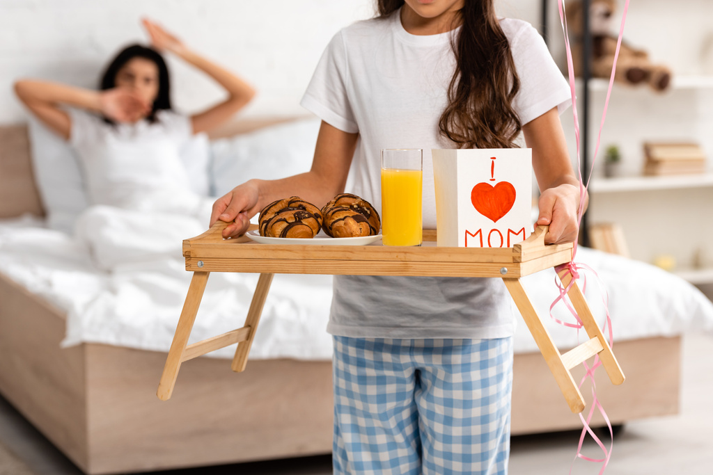 καλλιεργημένη άποψη του παιδιού κρατώντας δίσκο με πρωινό, κάρτα ημέρα μητέρες με την καρδιά σημάδι και γράμματα μαμά, ενώ η μητέρα τεντώνεται στο κρεβάτι - Φωτογραφία, εικόνα
