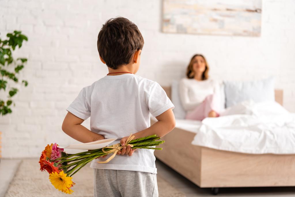 pohled zpět chlapec drží květiny a matky den karty, zatímco maminka sedí v posteli  - Fotografie, Obrázek