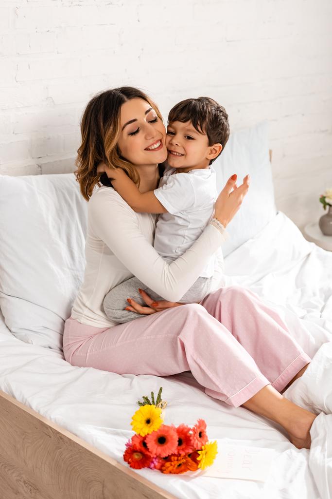счастливая женщина обнимает очаровательного сына, сидя в постели рядом с цветами в День матери
 - Фото, изображение