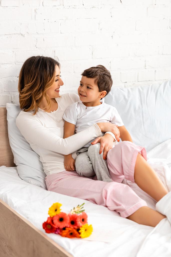 veselá maminka a syn při pohledu na sebe, zatímco sedí v posteli v blízkosti květin na den matek - Fotografie, Obrázek