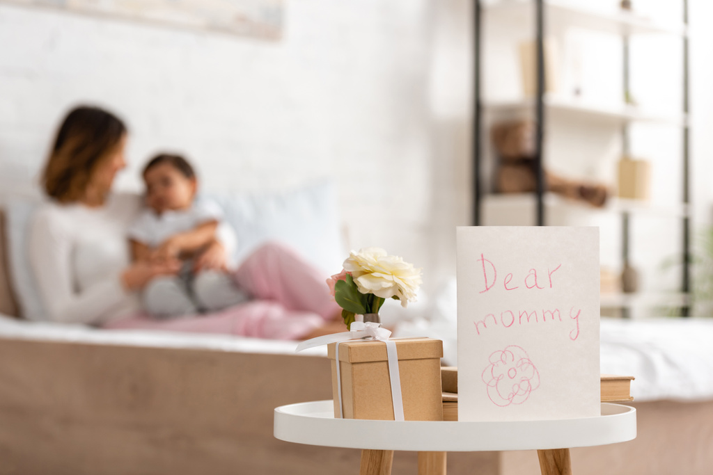 selektywne skupienie matczynej kartki dziennej z drogim napisem mamusi w pobliżu pudełka i róż na stole, i szczęśliwa kobieta siedząca na łóżku z synem  - Zdjęcie, obraz
