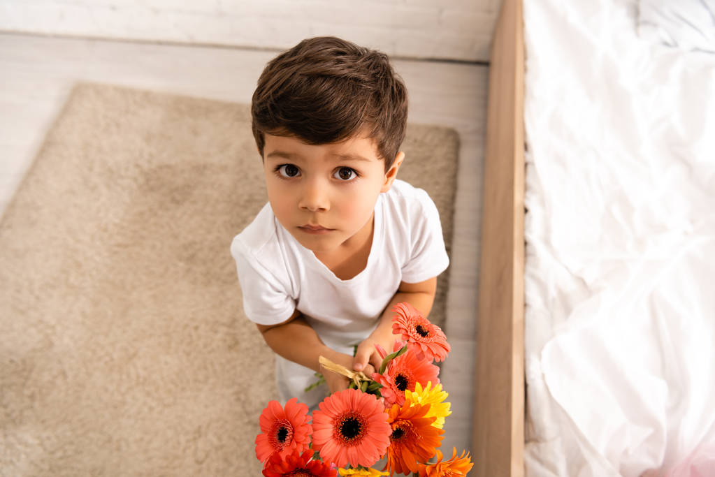 overhead view of adorable boy looking at camera while holding bouquet of gerberas on mothers day - Photo, Image