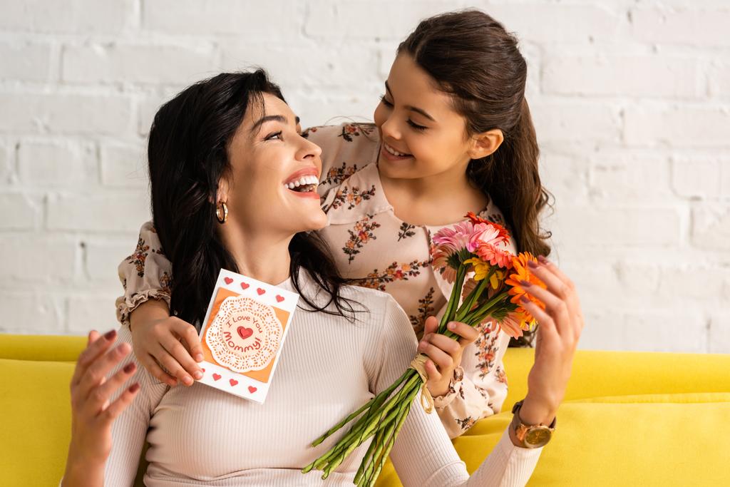 adorable hija sosteniendo madres tarjeta de día y flores mientras abraza feliz madre
 - Foto, imagen