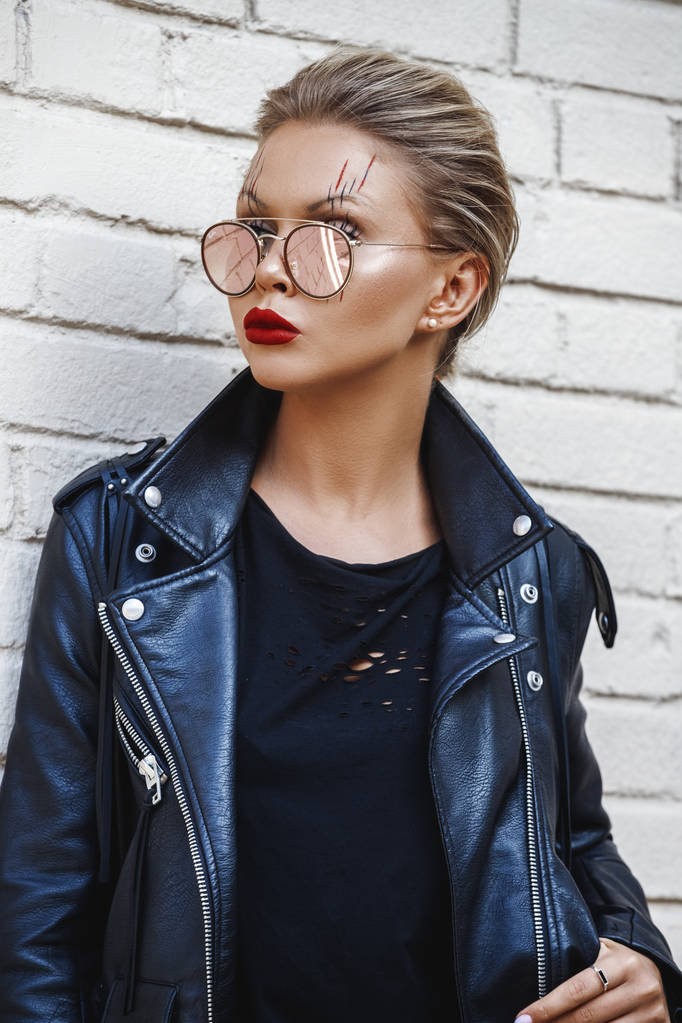 Beautiful blonde girl in black jacket and glasses posing on white brick wall background  - Photo, Image