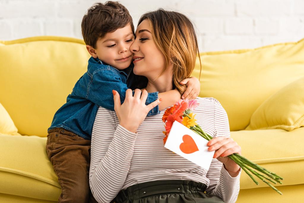 cute boy embracing mom holding flowers and mothers day card with heart symbol - Photo, Image