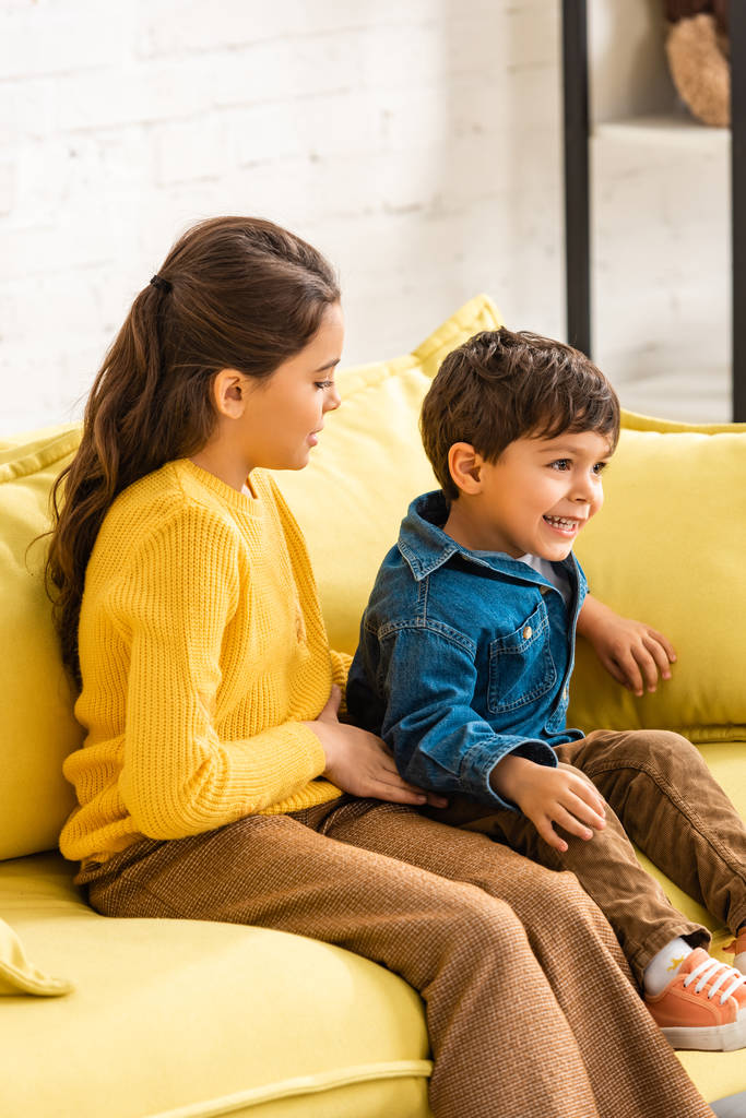 весёлый мальчик сидит на жёлтом диване рядом с очаровательной сестрой дома
 - Фото, изображение