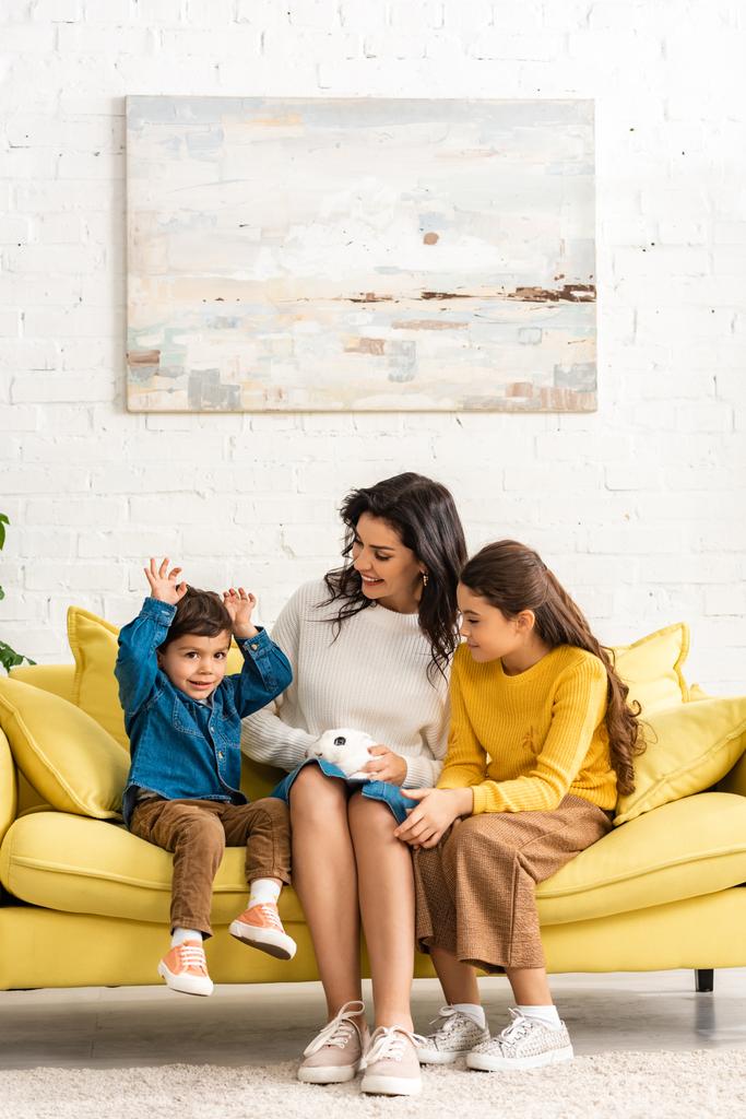 szczęśliwy dzieci i wesoły matka siedzi na kanapie z białym zabawnym królikiem - Zdjęcie, obraz
