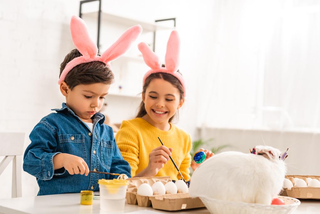 Tavşan kulaklı neşeli çocuklar paskalya yumurtalarını boyarken hasır şapkalı beyaz tavşanın yanında oturuyorlar. - Fotoğraf, Görsel