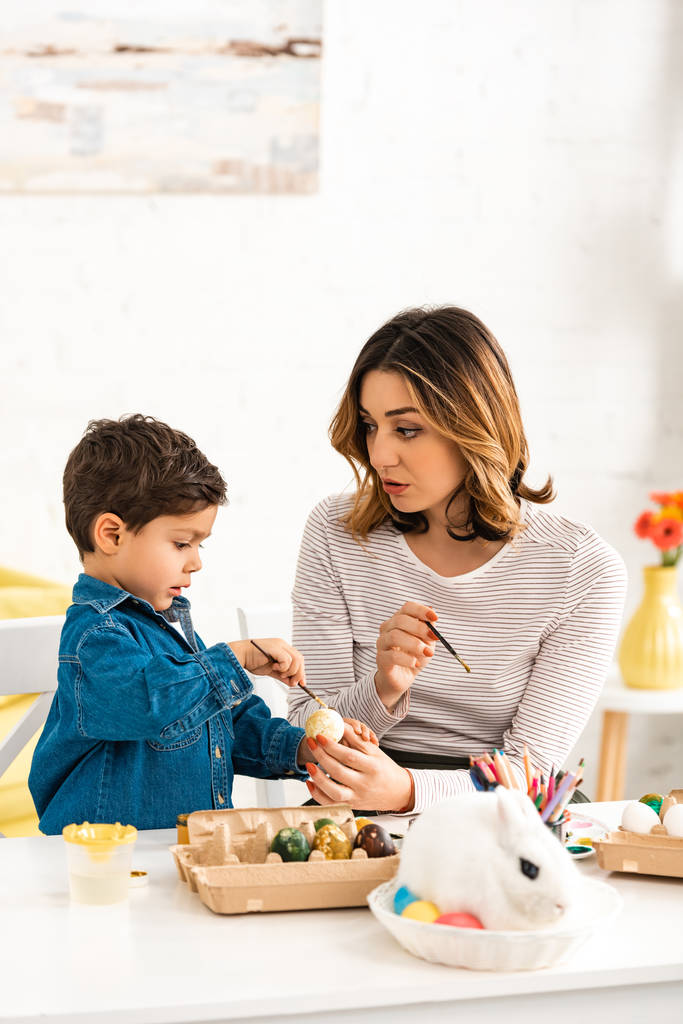 figyelmes anya és fia festés húsvéti tojás, miközben ül asztalnál közelében nyuszi fonott - Fotó, kép