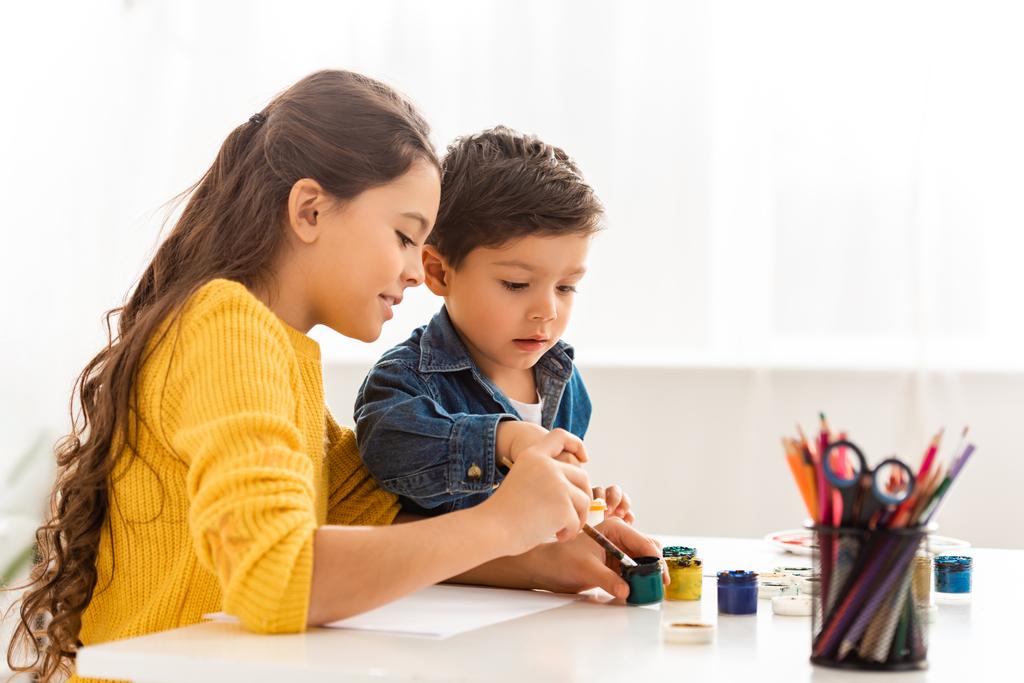 soustředěný bratr a sestra sedí u stolu a kreslí spolu s barvami - Fotografie, Obrázek