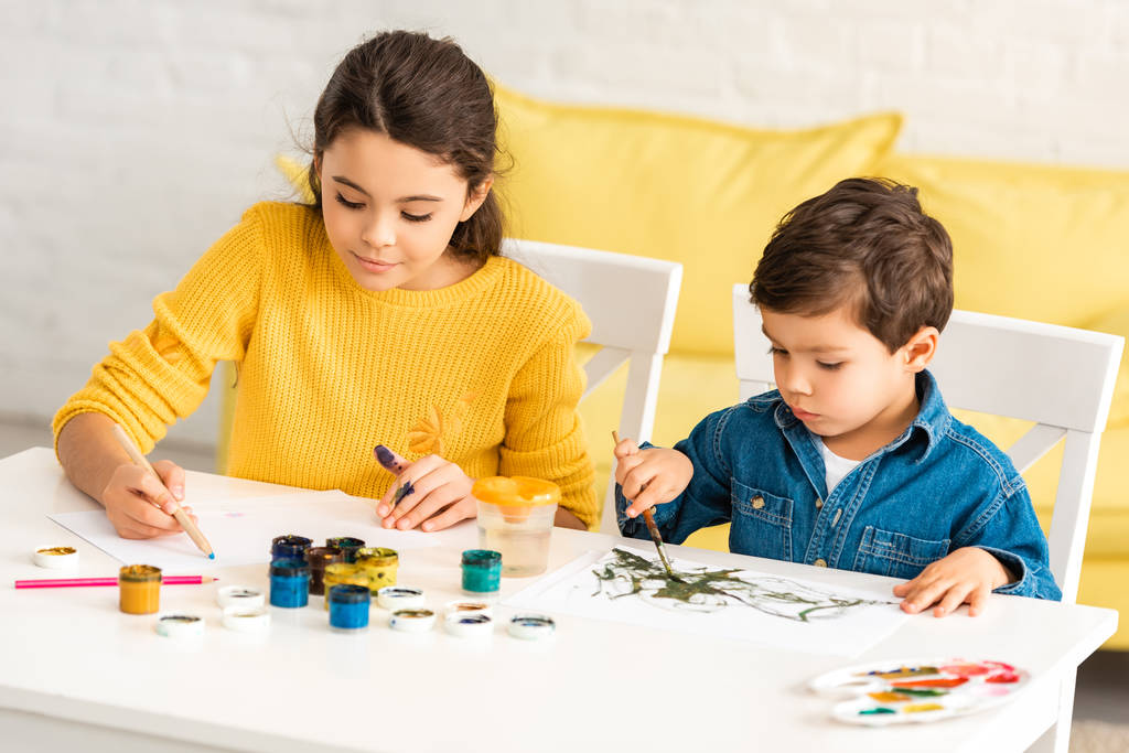 niños atentos y adorables sentados a la mesa y dibujando con pinturas juntos
 - Foto, Imagen