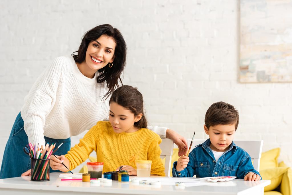 happy woman standing near desk and smiling at camera while adorable kids drawing with paints - Photo, Image