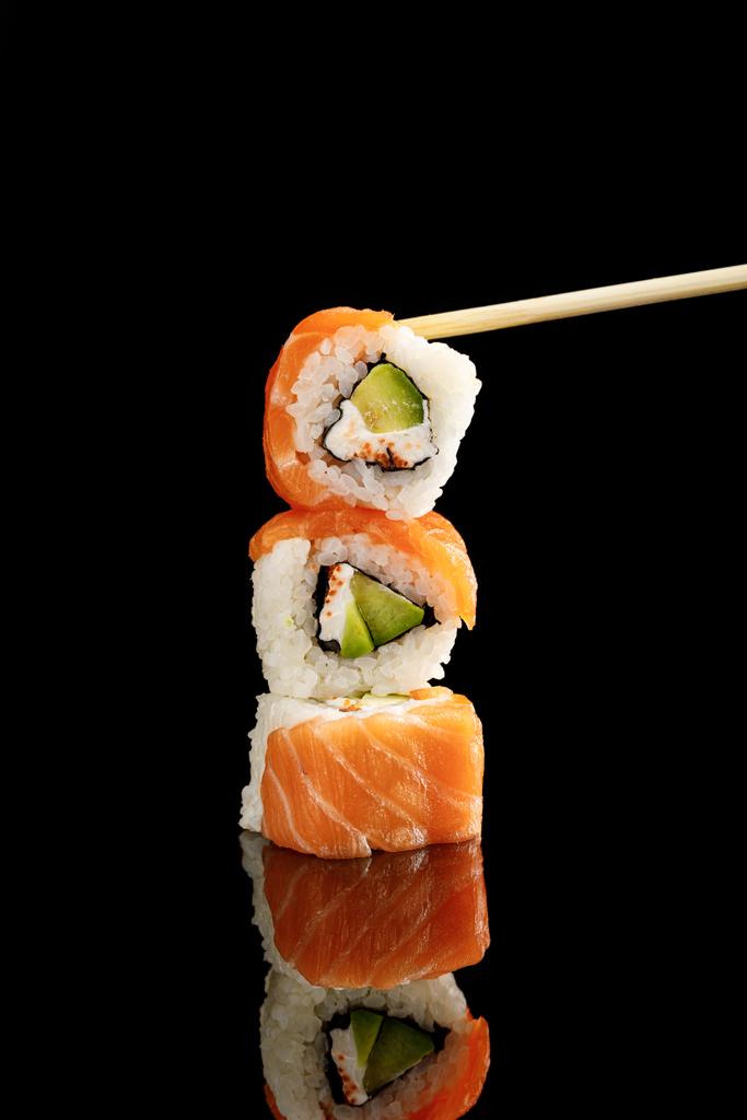 chopstick perto de delicioso sushi Filadélfia com abacate, queijo cremoso, salmão e masago caviar isolado em preto com reflexão
 - Foto, Imagem