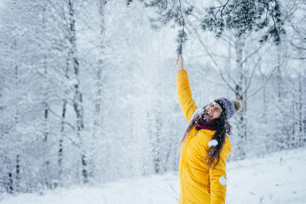 Młoda dziewczyna w dół kurtka i kapelusz w zimie las potrząsa śnieżną gałązką. - Zdjęcie, obraz