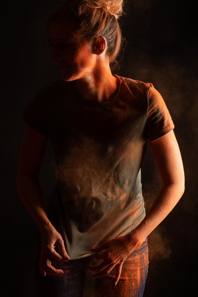 vista recortada de la mujer con polvo de pintura holi de color naranja en la ropa sobre fondo negro
 - Foto, Imagen