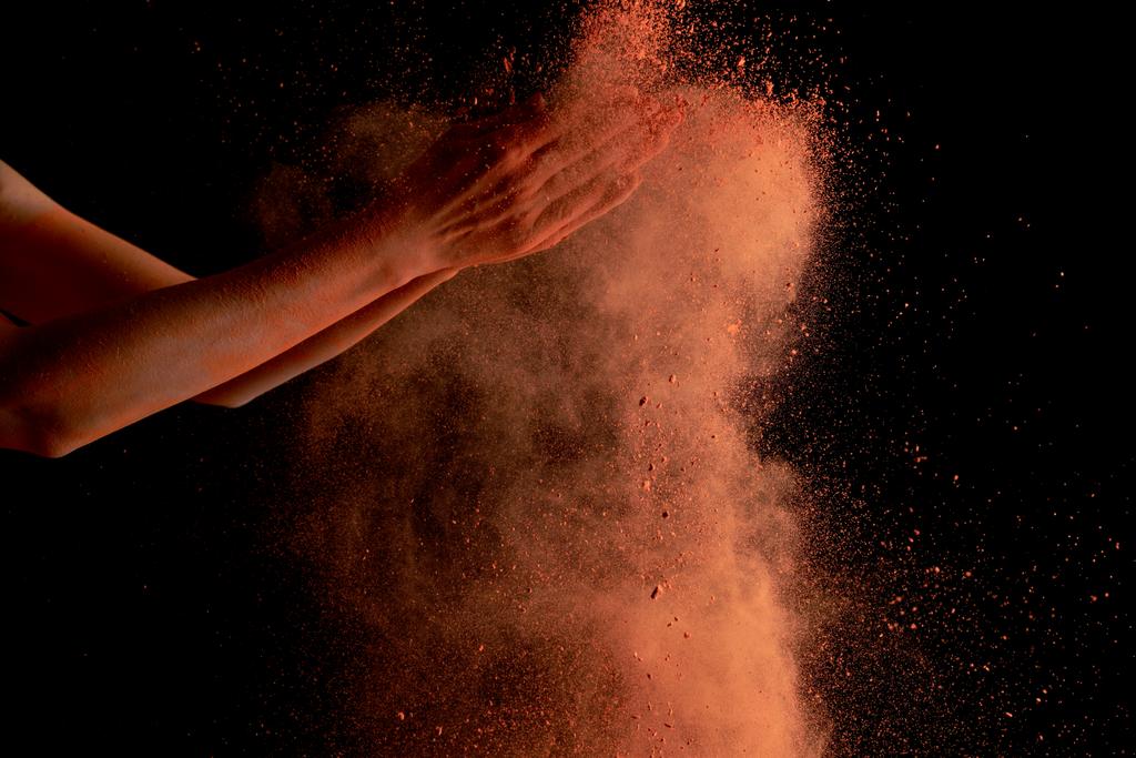 περικοπή άποψη των γυναικείων χεριών με πορτοκαλί πολύχρωμο χρώμα holi σκάσει σε μαύρο φόντο - Φωτογραφία, εικόνα