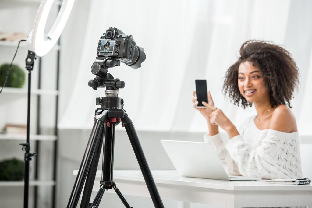 επιλεκτική εστίαση της ψηφιακής φωτογραφικής μηχανής κοντά σε ελκυστική αφρικανή blogger βίντεο σε τιράντες δείχνοντας με το δάχτυλο το smartphone με λευκή οθόνη  - Φωτογραφία, εικόνα