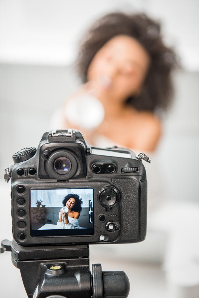 selectieve focus van digitale camera met krullend Afrikaanse Amerikaanse influencer in beugels houden container met cosmetische crème op display  - Foto, afbeelding