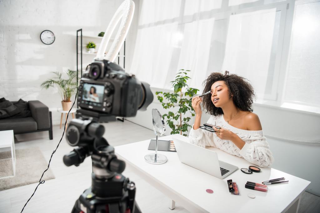 selective focus of curly african american influencer in braces applying makeup near digital camera  - 写真・画像
