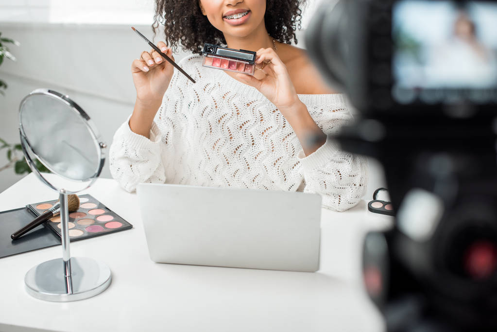 cropped view of happy african american video blogger in braces holding palette with lip gloss near digital camera  - Photo, Image