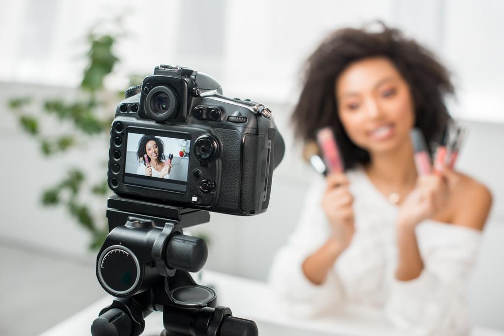 foco seletivo da câmera digital com curly blogueiro de vídeo americano africano em aparelho segurando gloss labial em exibição
  - Foto, Imagem