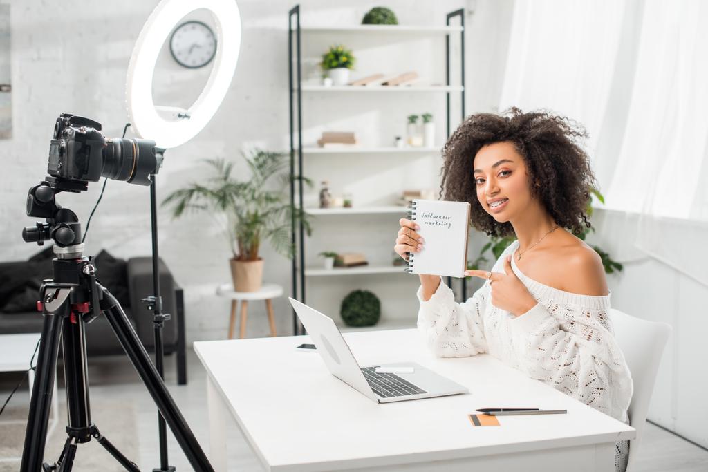 happy african american video blogger in braces pointing with finger at notebook with influencer marketing lettering near digital camera  - Photo, Image