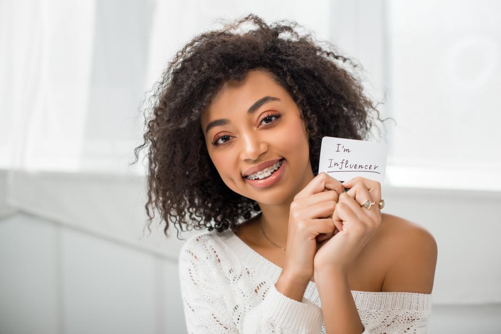 alegre afroamericana chica en frenos celebración tarjeta con yo soy influencer lettering en casa
  - Foto, imagen